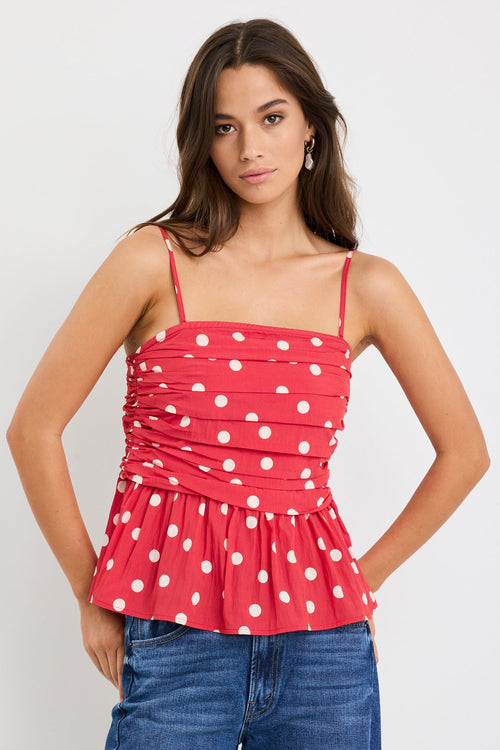 Woman wearing a red polka dot ruched cami top with thin straps and a peplum hem, styled with blue jeans against a plain white background.