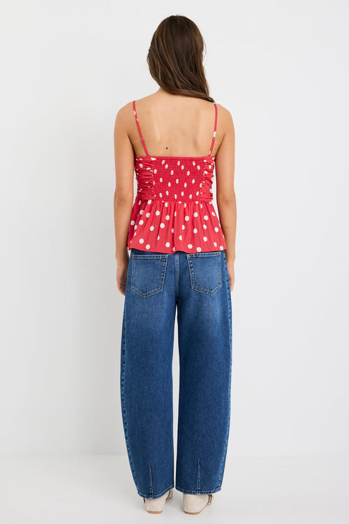 Woman wearing a red polka dot ruched cami top with thin straps and a peplum hem, styled with blue jeans against a plain white background.