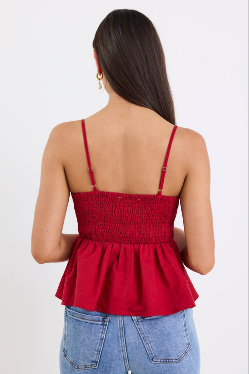 Woman wearing a plum-red pleated bodice peplum top with thin straps, paired with blue jeans and gold jewellery, standing against a white background.