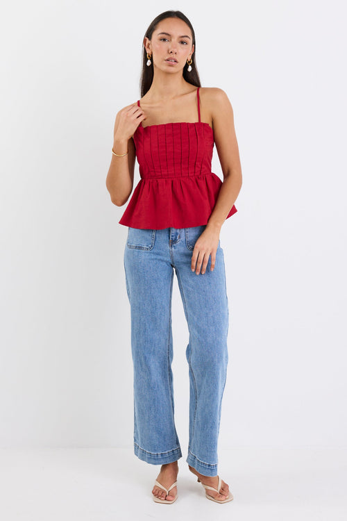 Woman wearing a plum-red pleated bodice peplum top with thin straps, paired with blue jeans and gold jewellery, standing against a white background.