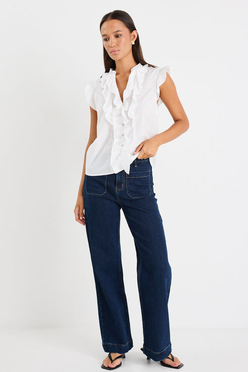 Woman wearing a sleeveless white blouse with ruffle detailing down the front and mixed buttons, paired with dark denim jeans.