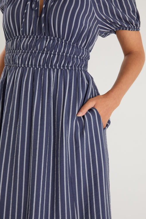 Woman wearing a navy maxi dress with vertical white stripes, short puff sleeves, a smocked waist, and side slit detail.