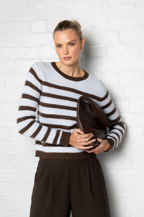 Woman wearing a brown and white striped sweater holding a clutch against a white brick wall.
