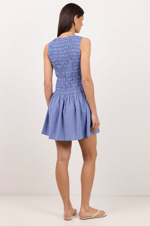 A woman is shown from the back wearing a sleeveless blue and white gingham mini dress with a smocked bodice and flared skirt, standing against a plain white studio background.