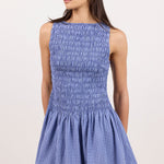 A woman modeling a sleeveless smocked mini dress with a blue and white gingham pattern, photographed against a plain light beige studio background.