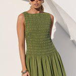 A woman modeling a sleeveless olive green dress with a smocked bodice and flared skirt, standing against a white wall with architectural shadows.