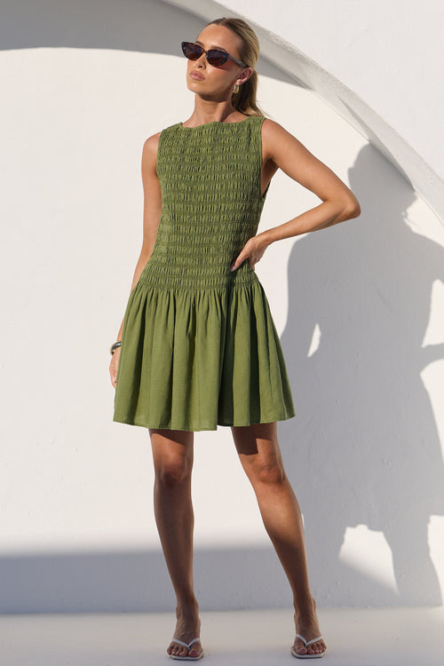 A woman modeling a sleeveless olive green mini dress with a smocked bodice and flared skirt, standing in front of a white wall with a strong shadow.