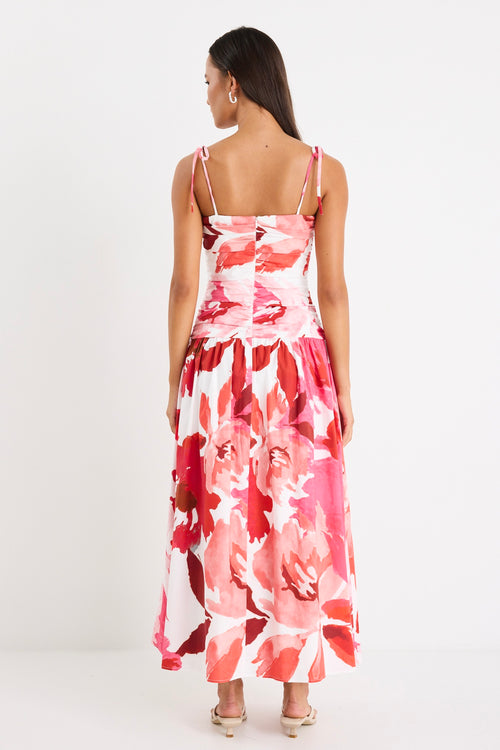 Woman wearing a floral pink and red ruched midi dress with tie-up straps and a flared hem, styled with minimal sandals.