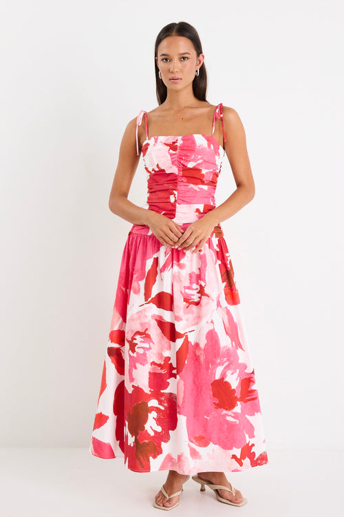 Woman wearing a floral pink and red ruched midi dress with tie-up straps and a flared hem, styled with minimal sandals.