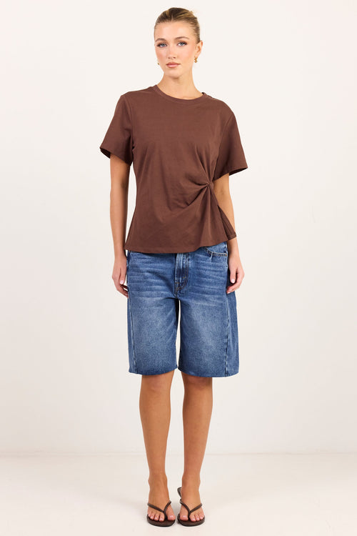 A woman modeling a brown short-sleeve t-shirt with a twisted side detail and blue denim shorts against a plain white studio background.