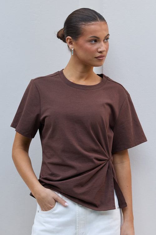 A woman wearing a brown short-sleeve t-shirt with a twisted knot detail at the hem, paired with white pants, standing against a light gray wall.
