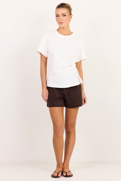 A woman modeling a white short-sleeve wrap-style t-shirt and dark brown shorts against a plain white studio background.
