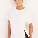 A woman modeling a white short-sleeve t-shirt with a twisted knot detail at the side hem, paired with dark brown pants, against a plain white background.