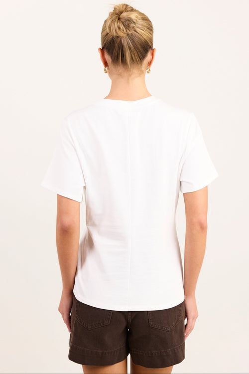A woman viewed from behind wearing a plain white short-sleeve t-shirt and dark brown shorts against a light off-white background.