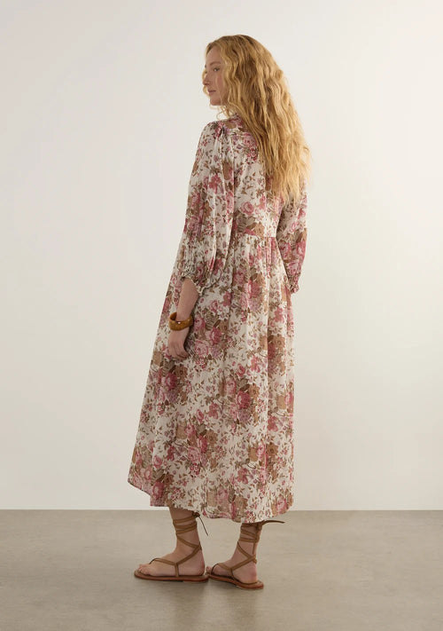 A woman with long wavy blonde hair is shown from the back wearing a long-sleeved midi dress with a pink and green floral pattern on a cream background, paired with brown strappy flat sandals, in a minimalist studio setting.