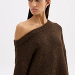 A woman modeling a dark brown, chunky knit off-the-shoulder sweater against a plain light gray studio background.