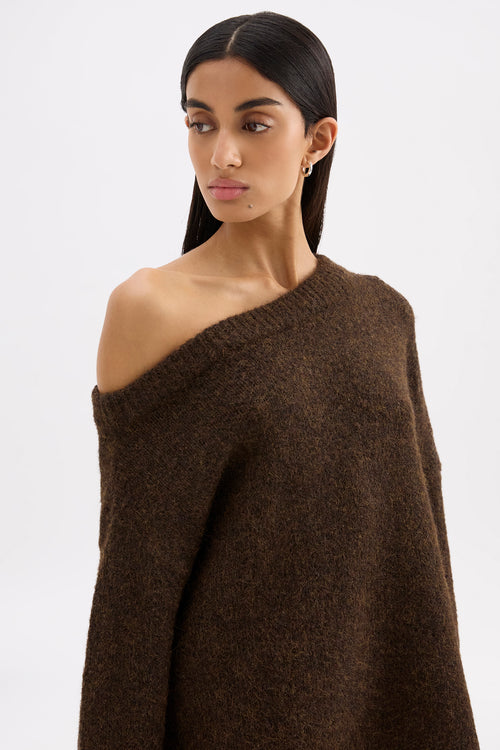 A woman modeling a dark brown, chunky knit off-the-shoulder sweater against a plain light gray studio background.