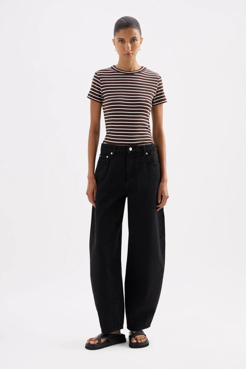 A model stands against a light gray studio background wearing a short-sleeve crewneck t-shirt with thin brown and white horizontal stripes, paired with black wide-leg jeans and black slide sandals.