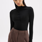 A woman modeling a black long-sleeve mock neck top paired with brown trousers, shot against a plain light gray studio background.