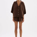 A model stands against a light gray studio background wearing a matching dark brown short-sleeve button-front shirt and shorts set, with black slide sandals.