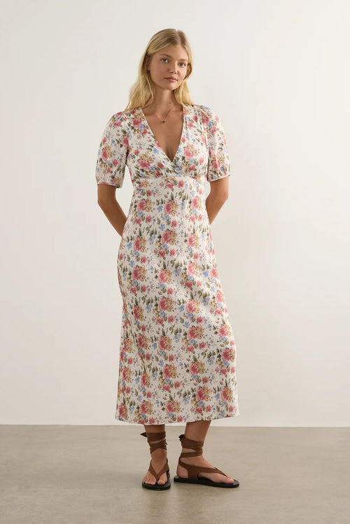 A woman modeling a short-sleeve midi dress with a pink, red, and green floral pattern on a cream background, paired with brown strappy sandals, against a plain light gray studio backdrop.