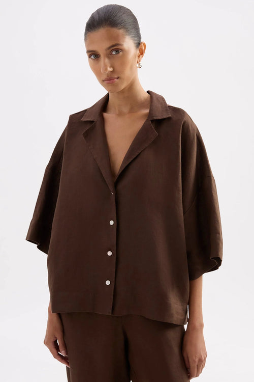 A woman wearing a dark brown short-sleeve button-up shirt and matching brown trousers, photographed against a plain light gray studio background.