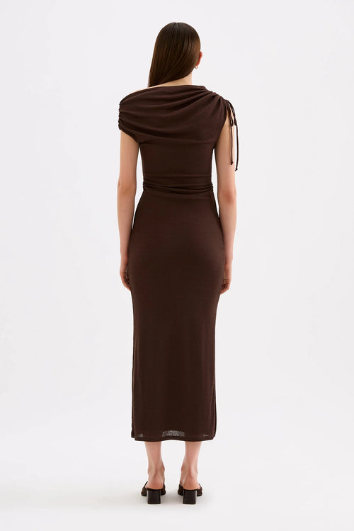 A woman viewed from the back wearing a dark brown draped midi dress with short sleeves and side tie details, paired with black heels, against a plain white studio background.