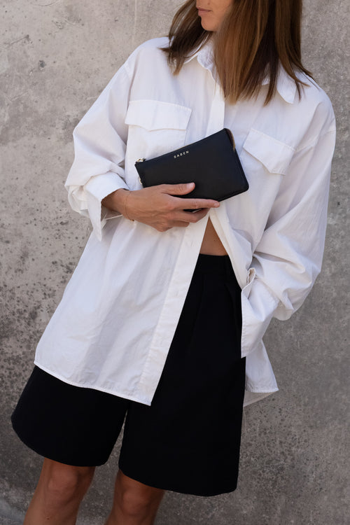 A woman wearing an oversized white button-up shirt and black shorts, holding a black leather clutch bag with 'SUEYU' printed on the front, standing against a textured gray concrete wall.