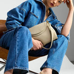 A model sits on a wooden chair wearing a blue denim jacket and matching wide-leg jeans, holding a beige shoulder bag, and wearing beige slingback heels against a studio background.