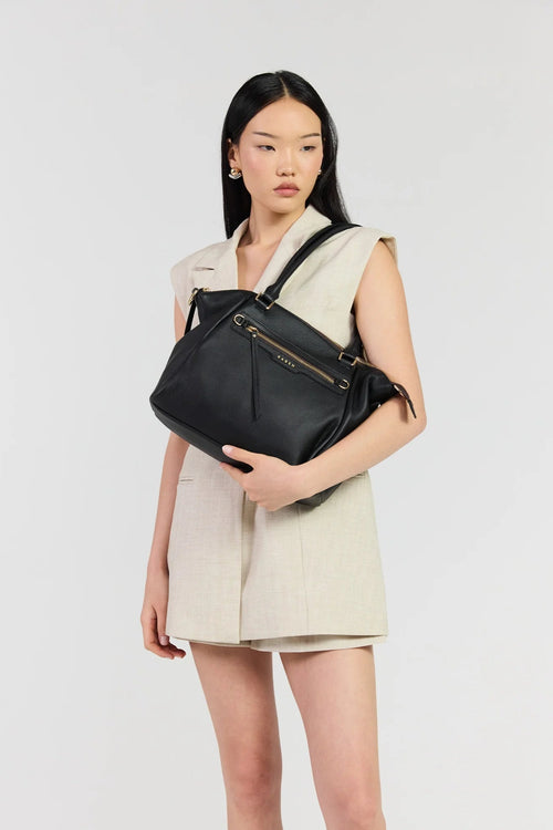 A model wearing a beige sleeveless blazer dress holds a black leather shoulder bag with gold hardware against a light gray studio background.