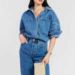 A model wearing a blue denim shirt-jacket and matching blue denim jeans, holding a beige clutch bag against a light gray studio background.