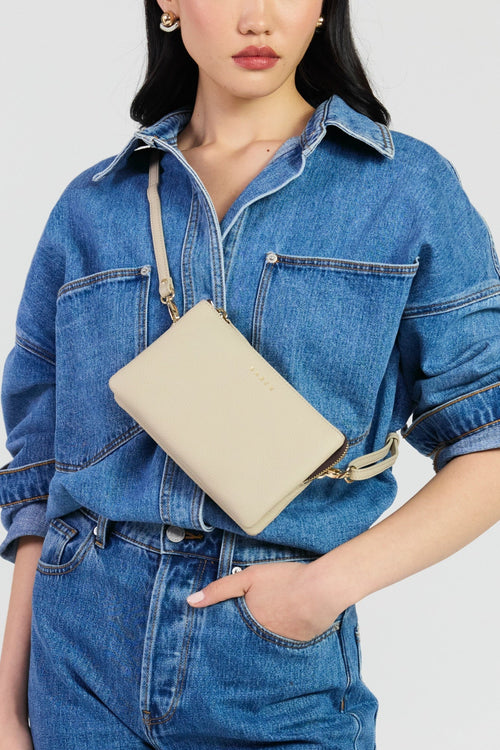 A woman wearing a blue denim shirt and matching denim pants carries a beige leather crossbody bag with a matching strap and gold-tone hardware against a light gray studio background.