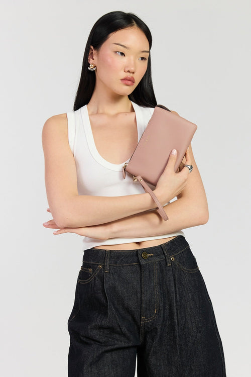 A woman with long black hair models a dusty pink leather clutch bag with a wristlet strap, worn over a white tank top and dark denim jeans, against a light gray studio background.