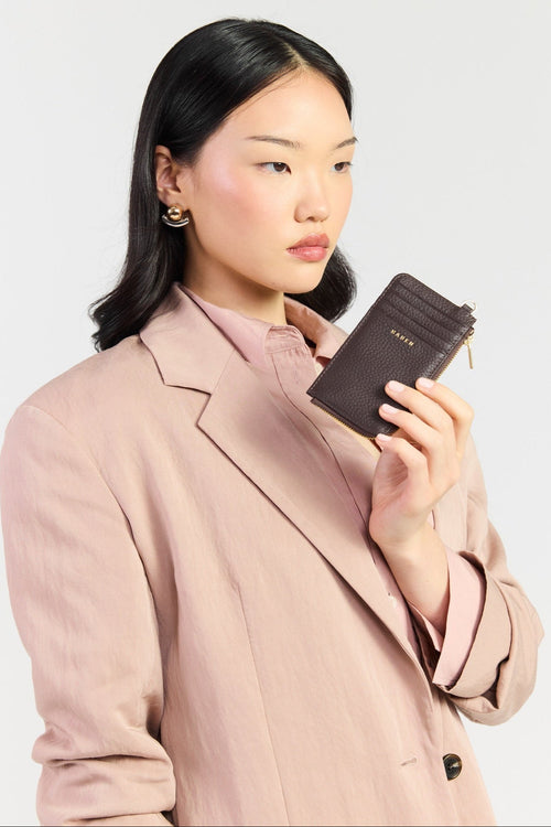 A woman wearing a light pink blazer holds a dark brown leather wallet with gold lettering that reads 'SARA' against a plain light gray background.