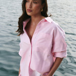 Woman in a pink shirt and white shorts standing by a body of water with boats in the background.