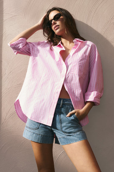 Woman wearing a pink shirt and denim shorts against a textured wall.
