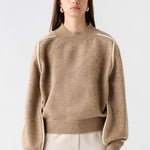 Woman wearing a beige sweater and white pants, holding a beige handbag on a white background