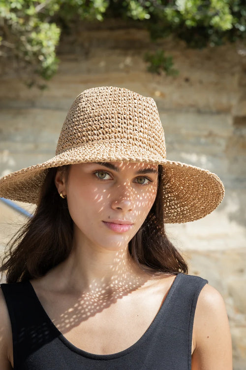A woman wearing a wide-brimmed woven straw sun hat with a black tank top, photographed outdoors against a stone wall with dappled sunlight and green foliage.