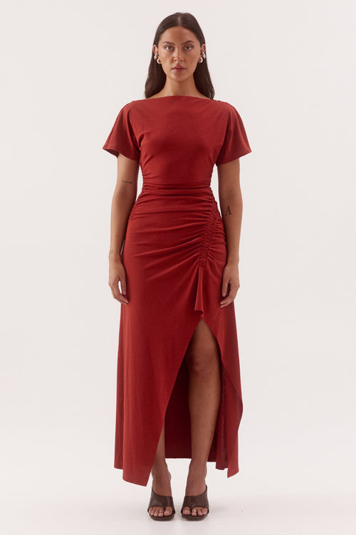A woman modeling a rust-red midi dress with short flutter sleeves, a boat neckline, a ruched waist, and a high-low front slit, photographed against a seamless white studio background.