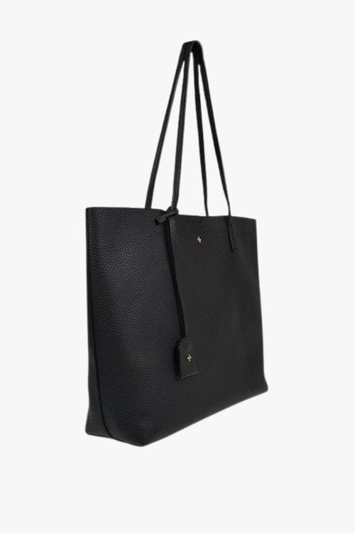 A sleek black pebble-textured vegan leather tote bag with long shoulder straps, minimal gold detailing, and a small matching tag charm hanging from one handle. The bag has a structured rectangular shape and a clean, minimalist design