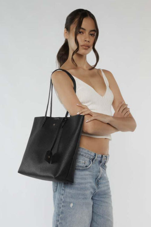 A sleek black pebble-textured vegan leather tote bag with long shoulder straps, minimal gold detailing, and a small matching tag charm hanging from one handle. The bag has a structured rectangular shape and a clean, minimalist design