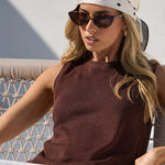 A woman sits in a wooden lounge chair outdoors, wearing a sleeveless dark brown top, white pants, a white bandana with black polka dots, and sunglasses.