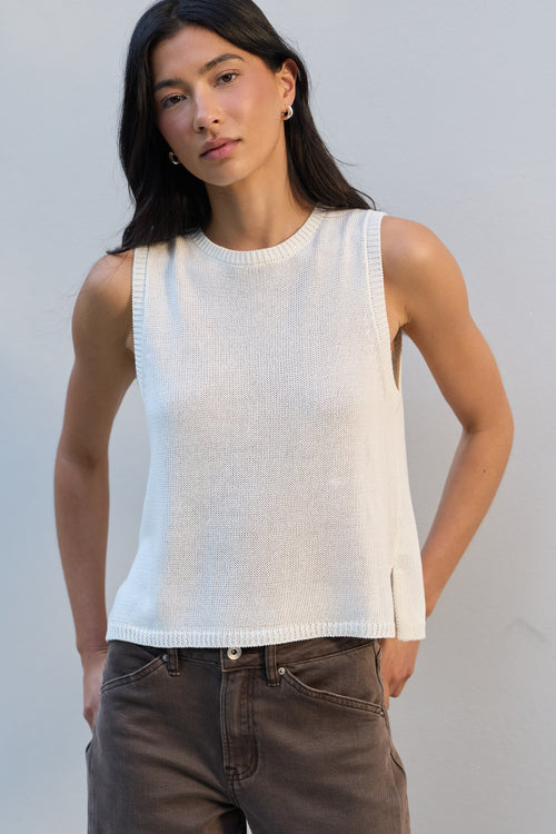 A woman wearing a sleeveless white knit top and brown jeans, standing against a plain light gray wall.