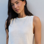 A woman with long dark hair wearing a ribbed white sleeveless knit top, posing against a light gray wall.