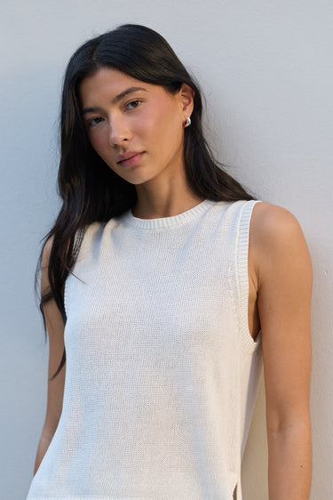 A woman with long dark hair wearing a ribbed white sleeveless knit top, posing against a light gray wall.