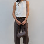 A woman stands against a light gray wall wearing a sleeveless white ribbed knit top, dark brown wide-leg pants, and brown sandals, while holding a taupe-brown crescent shoulder bag.