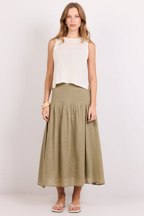 A woman stands against a plain light gray studio background wearing a sleeveless white top and a long olive green pleated skirt, with neutral sandals and bracelets.