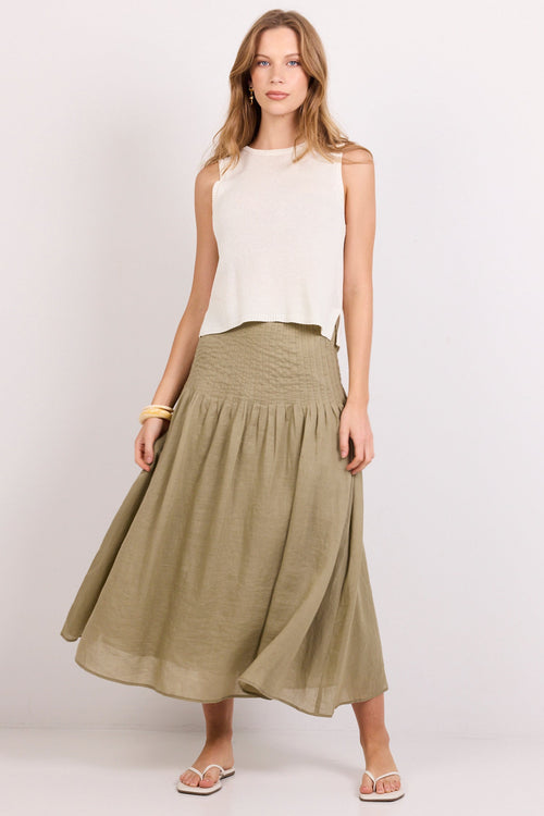 A woman modeling a sleeveless white top and a flowing olive green midi skirt against a plain light gray studio background.