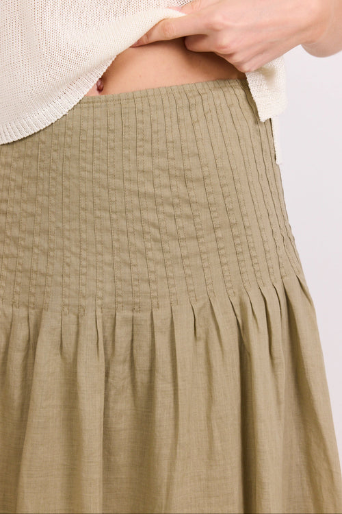 Close-up of a person wearing a textured sage green skirt with a smocked waistband and gathered pleated lower half, paired with a cream knit top against a light gray background.