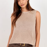 A woman modeling a sleeveless ribbed knit top in a light taupe-beige color, paired with dark brown pants and a gold belt, against a plain off-white studio background.